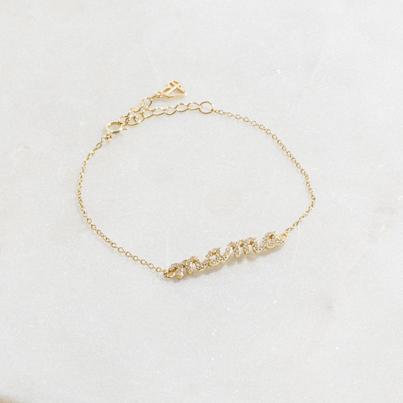 The Pave Mama Script Bracelet in gold, with "mama" spelled out in sparkling detail, rests on white marble—a stylish and elegant piece of jewelry perfect for celebrating motherhood.