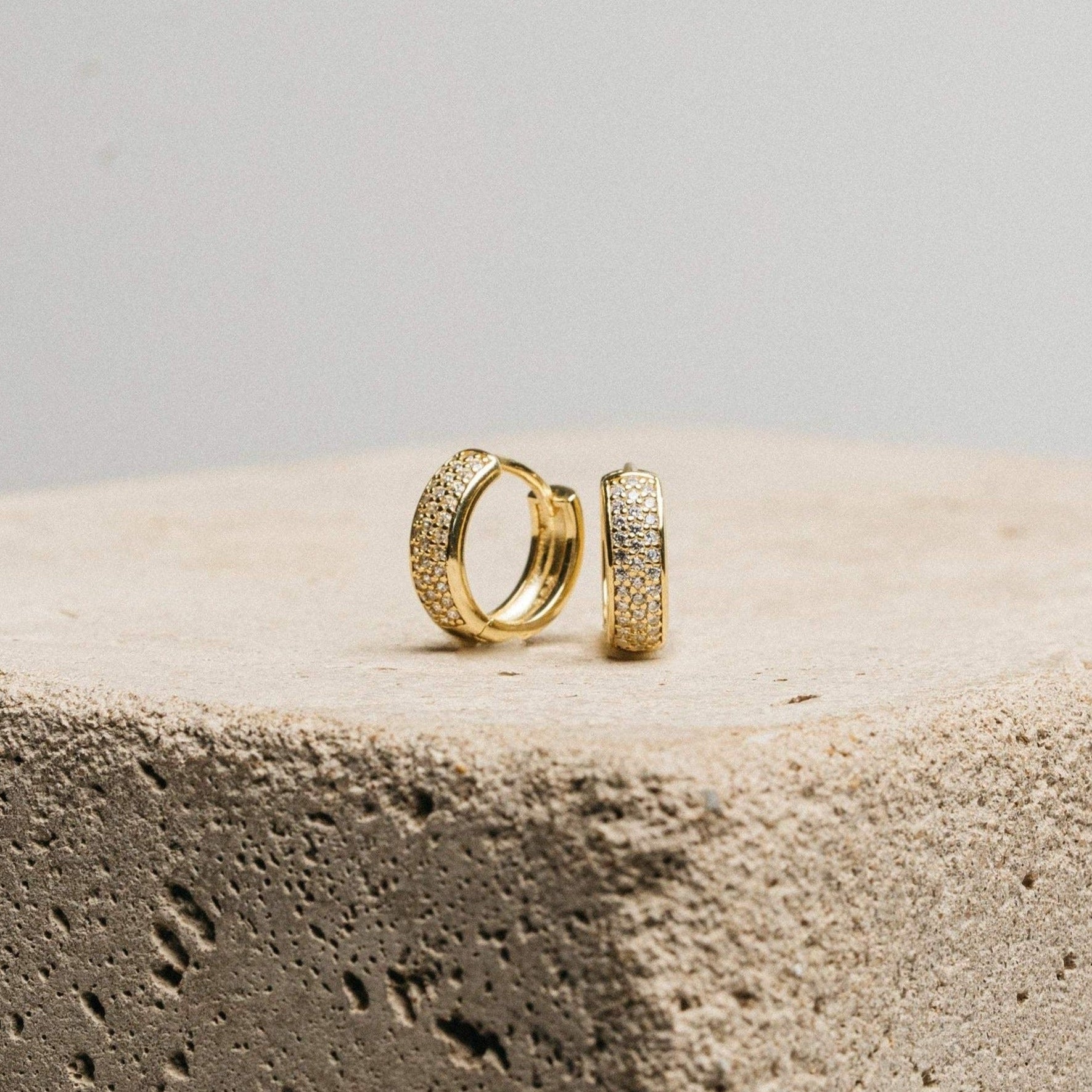 Pave Diamond Huggies rest on a textured stone block against a plain background—an elegant jewelry box essential, featuring sparkling diamond-encrusted hoops.