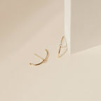 The Arc Studs, minimalist gold arcs with small diamonds, are shown on a light beige surface beside a block—ideal for fans of minimal ear jewelry.