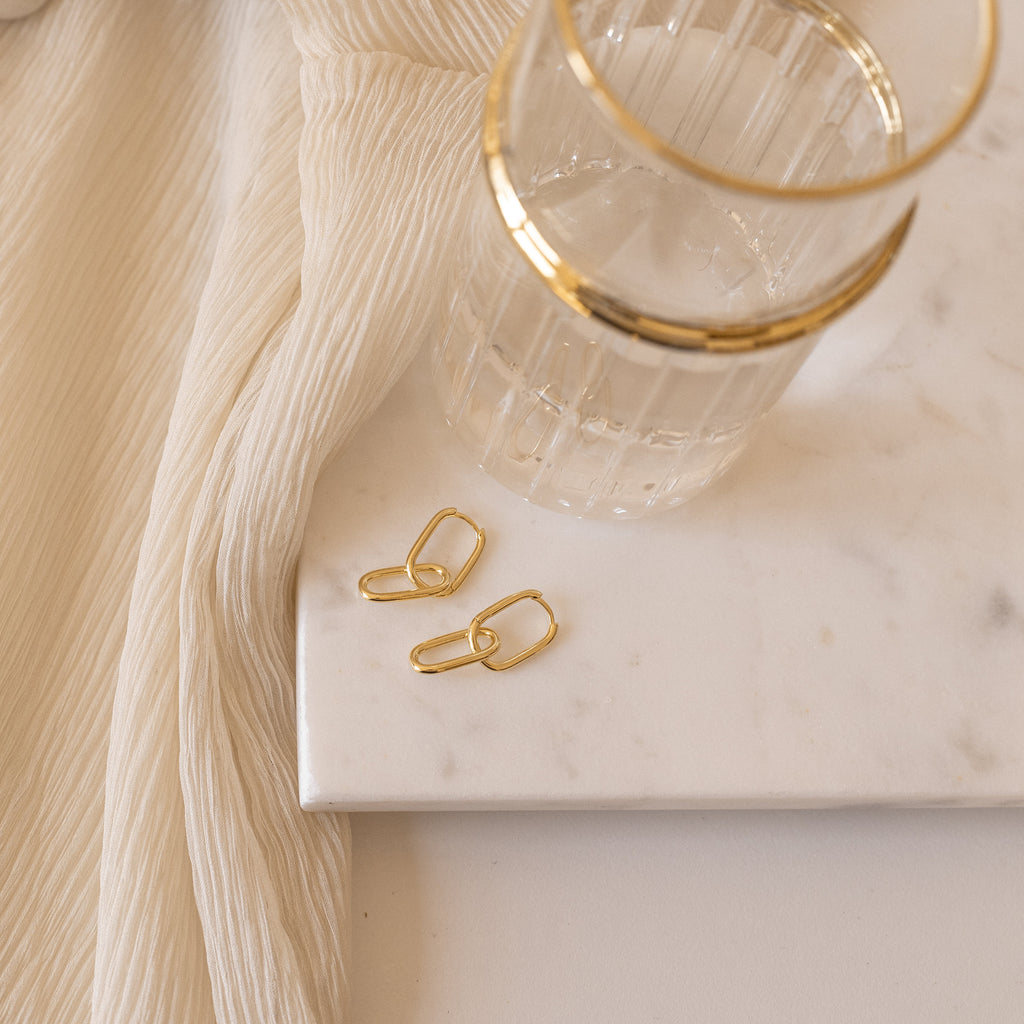 The Amanda Link Earrings in 18K Gold rest beside a glass of water on a marble tray, complemented by cream-colored fabric draped nearby.