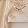 The Amanda Link Earrings in 18K Gold rest beside a glass of water on a marble tray, complemented by cream-colored fabric draped nearby.