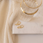 The Amanda Link Earrings in 18K Gold rest beside a glass of water on a marble tray, complemented by cream-colored fabric draped nearby.