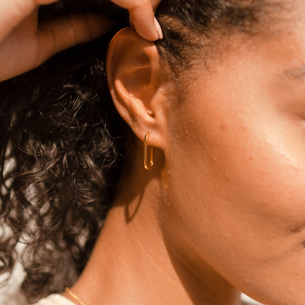 Close-up of a woman’s ear wearing Nina Link Earrings—gold geometric hoops—with curly hair visible in soft sunlight.