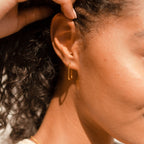 Close-up of a woman’s ear wearing Nina Link Earrings—gold geometric hoops—with curly hair visible in soft sunlight.