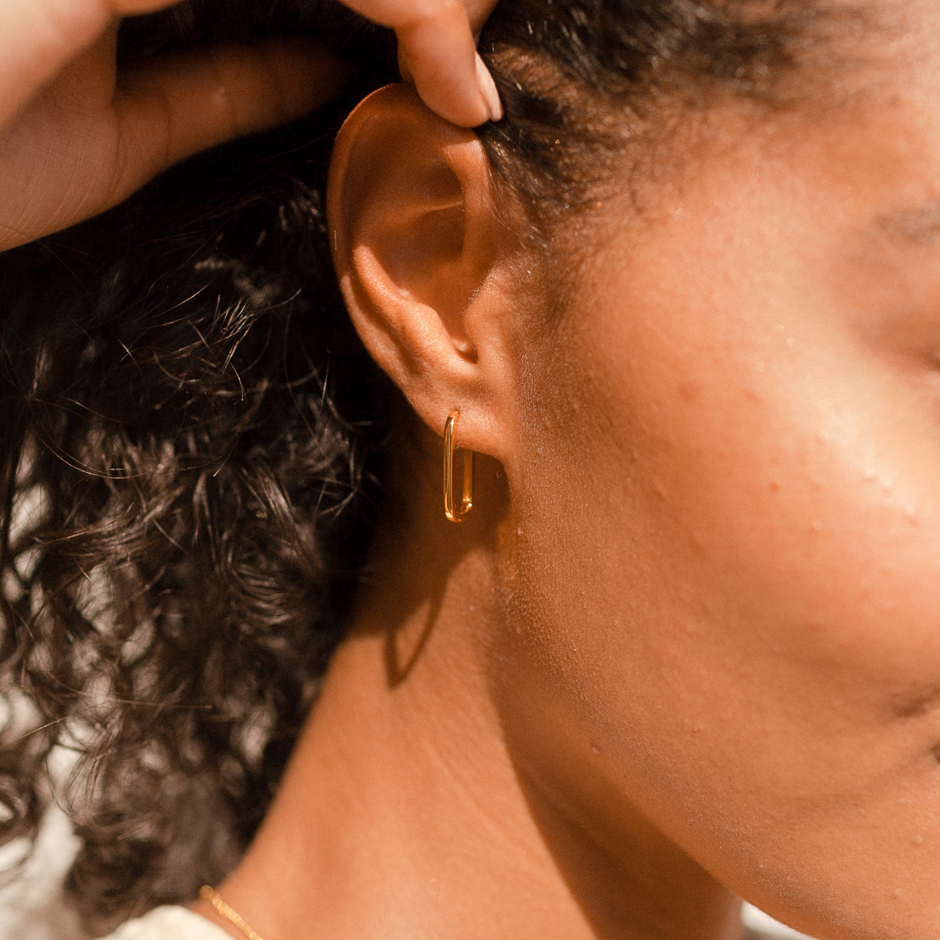 Close-up of a woman’s ear wearing Nina Link Earrings—gold geometric hoops—with curly hair visible in soft sunlight.