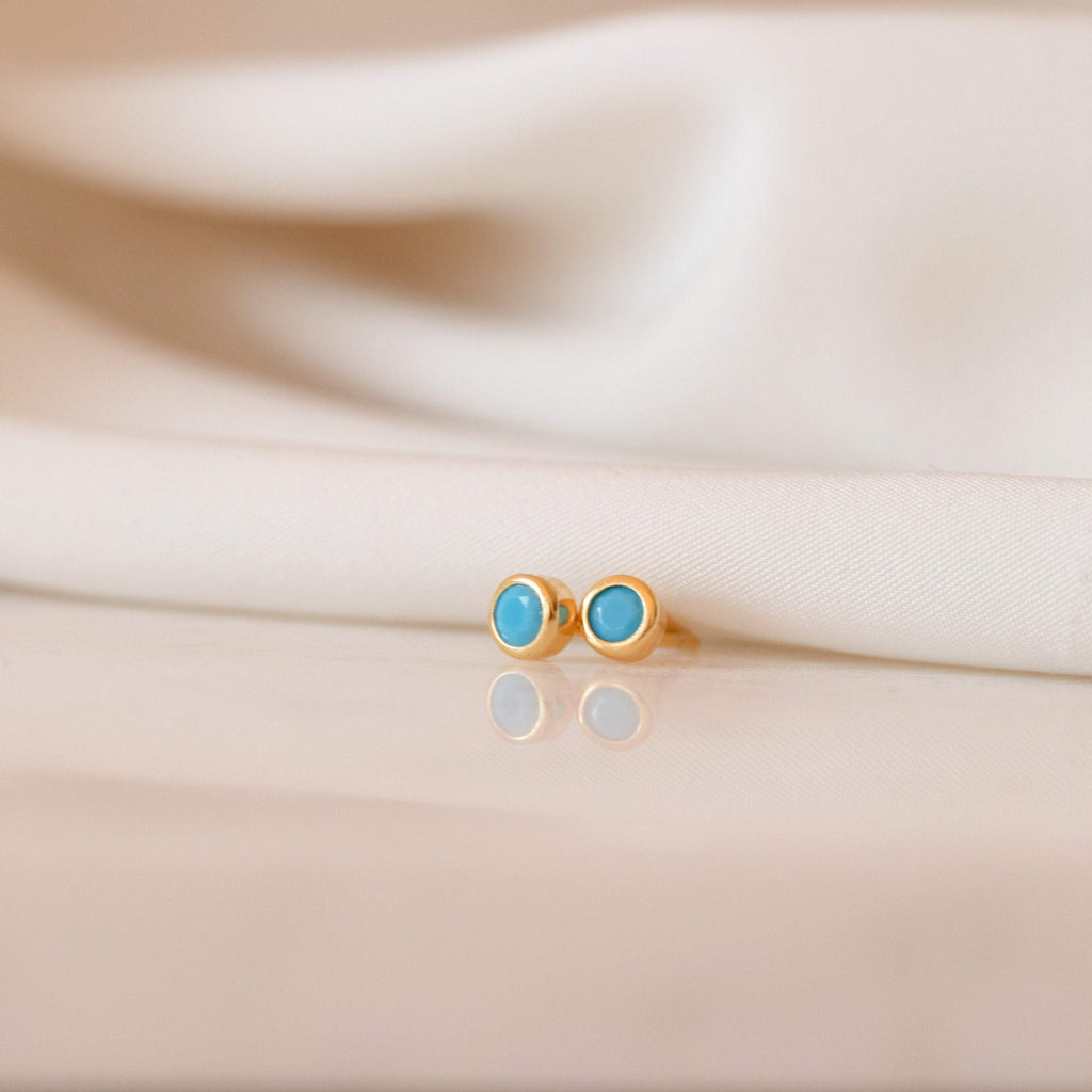 Ariel Turquoise Studs—dainty gold earrings with round turquoise stones—are elegantly displayed on white fabric, perfect for lovers of turquoise jewelry.