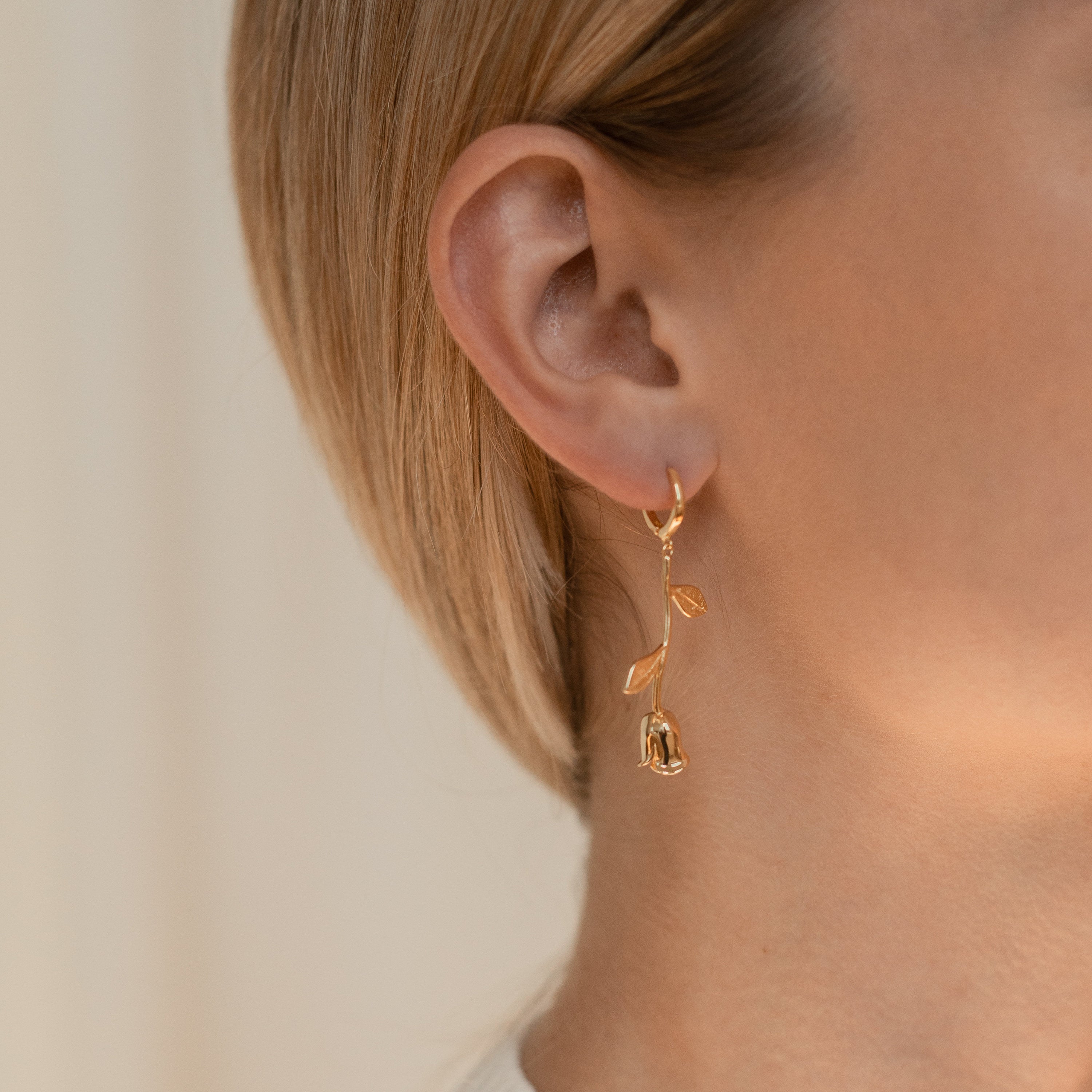 Close-up of a woman wearing Dangling Rose Huggies with her hair tucked behind her ear—a romantic and elegant gold rose-shaped earring.