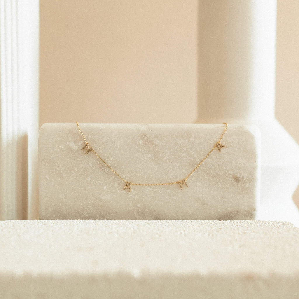 The Pave Mama Letter Necklace, shown with small charms on a marble block and neutral background, is a stylish piece of motherhood jewelry—making it the perfect gift for mom.