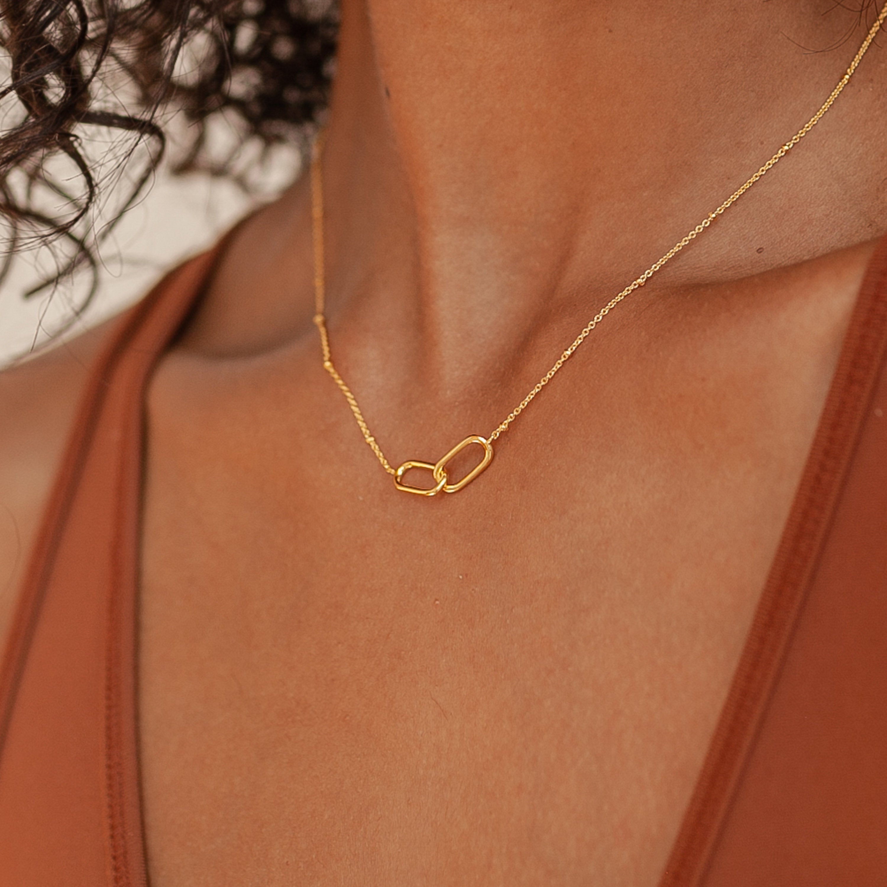 A person in a brown top wears the Linked Pendant Necklace—a thin gold chain featuring two interlocking loops symbolizing love.