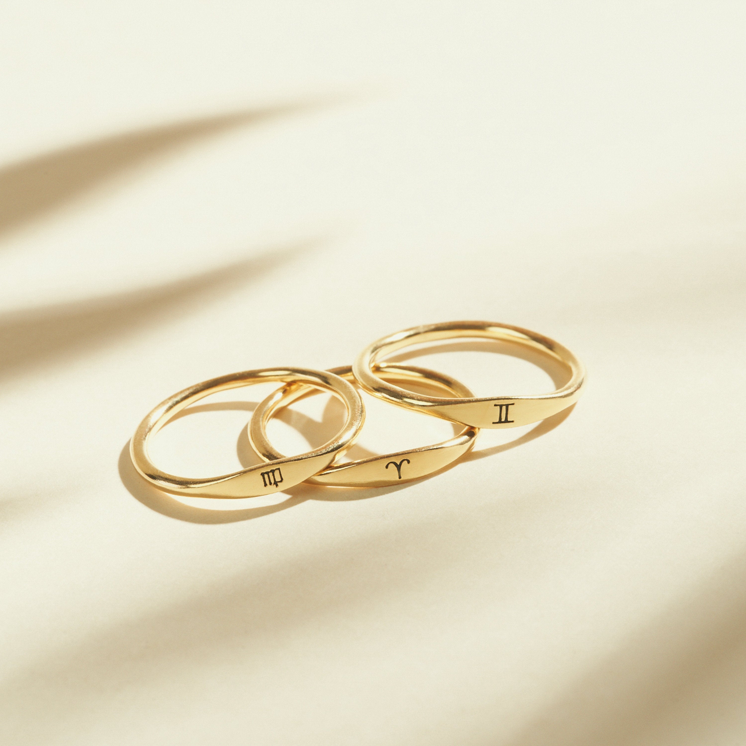 Three Skinny Zodiac Rings with engraved astrological signs rest on a light, neutral background with soft shadows—perfect stacking rings to add a personalized touch.