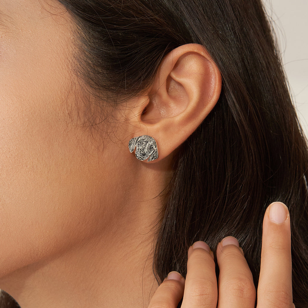 A close-up of a woman’s ear wearing Pet Portrait Studs, silver dog-shaped earrings perfect for pet lovers, with her hand near her face.