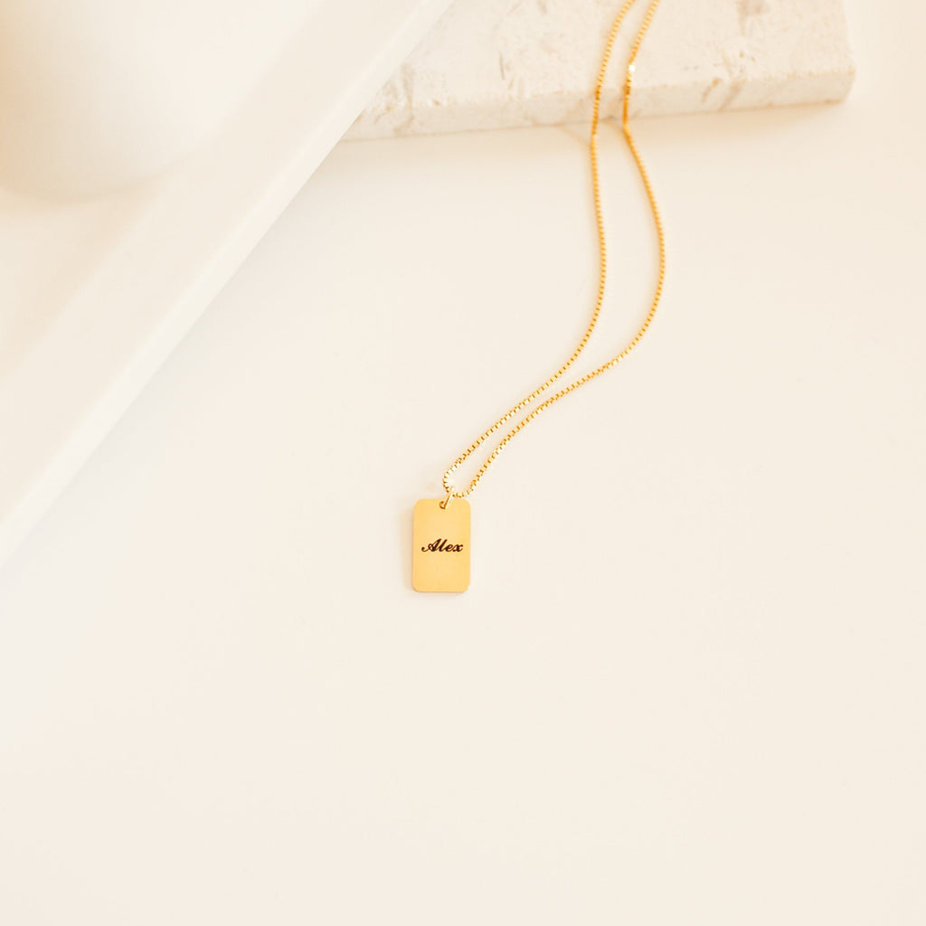 The Name Tag Necklace features a rectangular gold pendant engraved with Alex, displayed on a minimalist background—ideal for lovers of personalized jewelry.