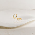 Two Cleo Cuffs, small gold hoop ear cuffs, rest on a wavy white ceramic dish against a neutral background, providing a stylish option for non-pierced earrings.
