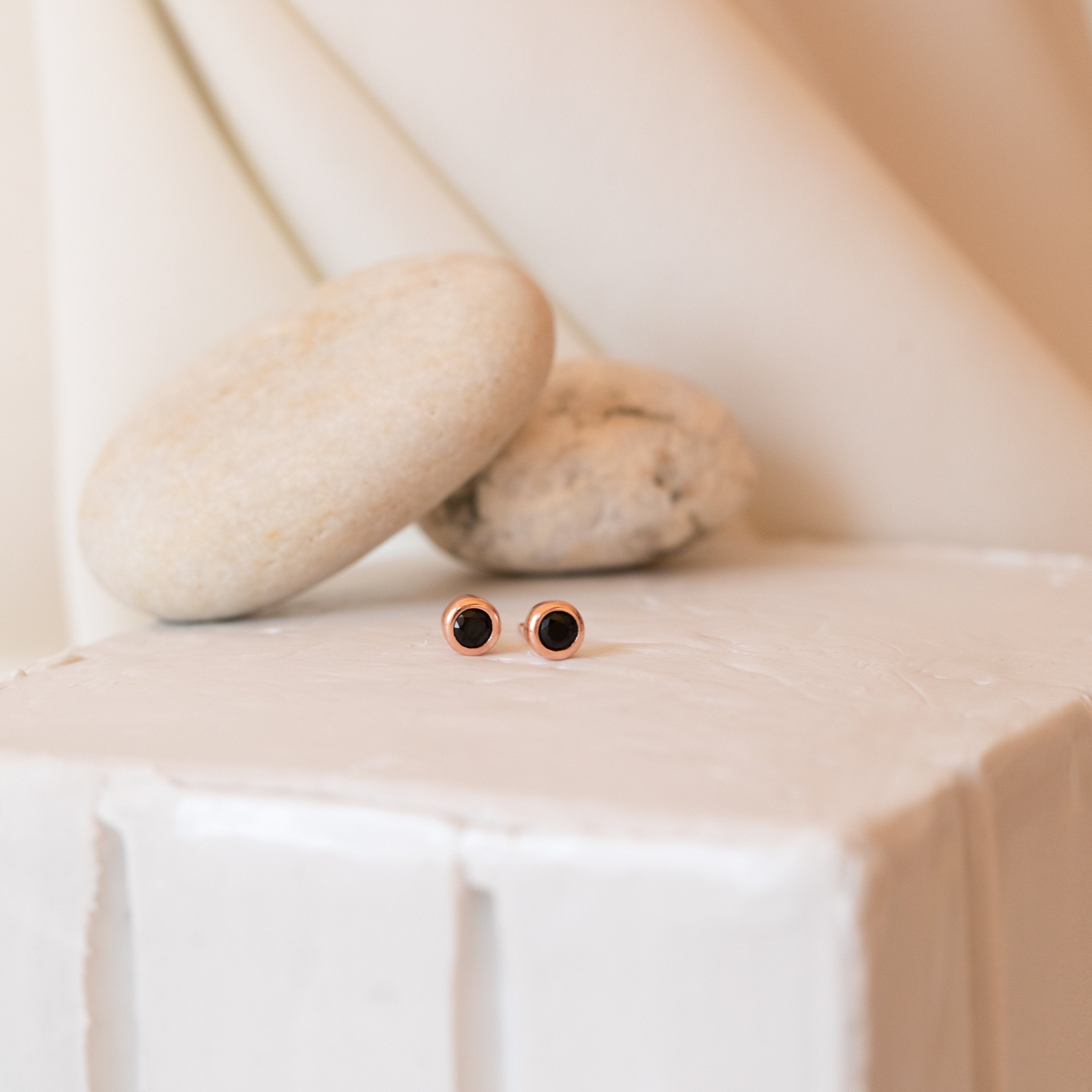 The Noir Diamond Studs feature small round black centers with gold edges, creating a unique look. Displayed on a white textured surface with decorative stones, these earrings make an elegant statement.