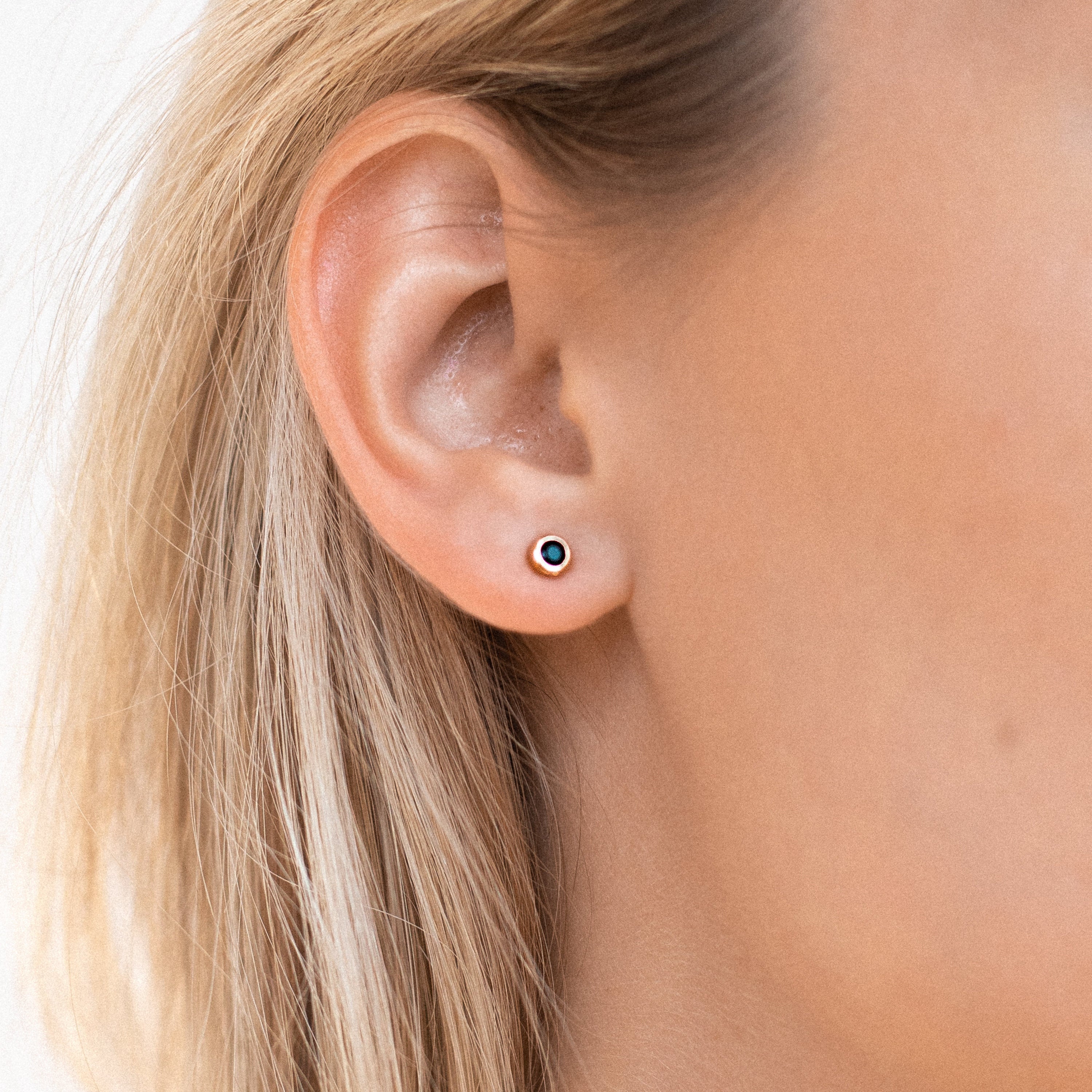 Close-up of a woman's ear with straight blonde hair, wearing Noir Diamond Studs—unique small round earrings featuring a dark center.