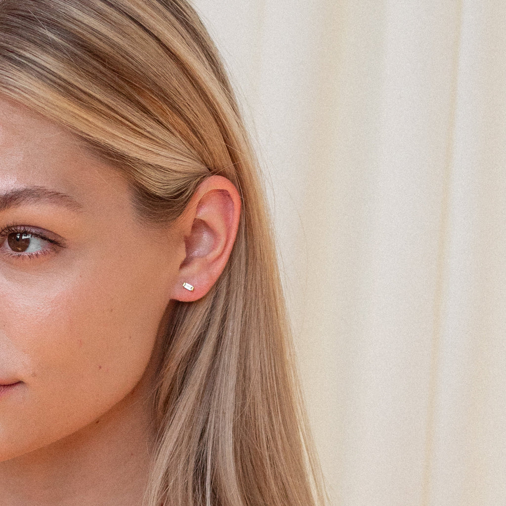 A woman's ear is shown wearing the Baguette Diamond Studs, with her blonde hair framing the elegant accessory against a light background.