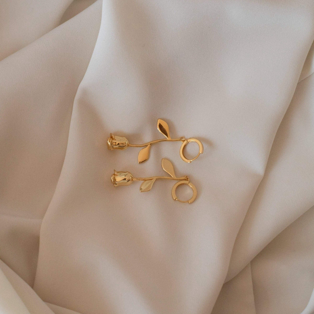 Dangling Rose Huggies—gold Venetian-inspired earrings shaped like roses with delicate leaves, displayed on soft ivory fabric. Ideal for anyone seeking romantic jewelry.