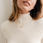 Woman in a cream ribbed turtleneck wearing the Pave Name Necklace, featuring sparkling pave diamonds; her face is partially visible.