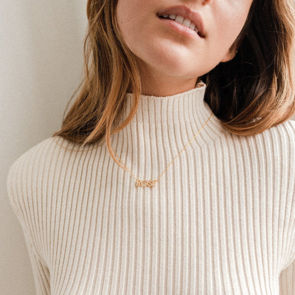 Woman in a cream ribbed turtleneck wearing the Pave Name Necklace, featuring sparkling pave diamonds; her face is partially visible.