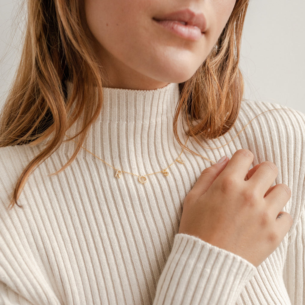 A woman in a cream ribbed sweater holds a custom Pave Letter Necklace in Rose Gold, featuring charms that spell ROSE —an ideal gift for fans of personalized jewelry.