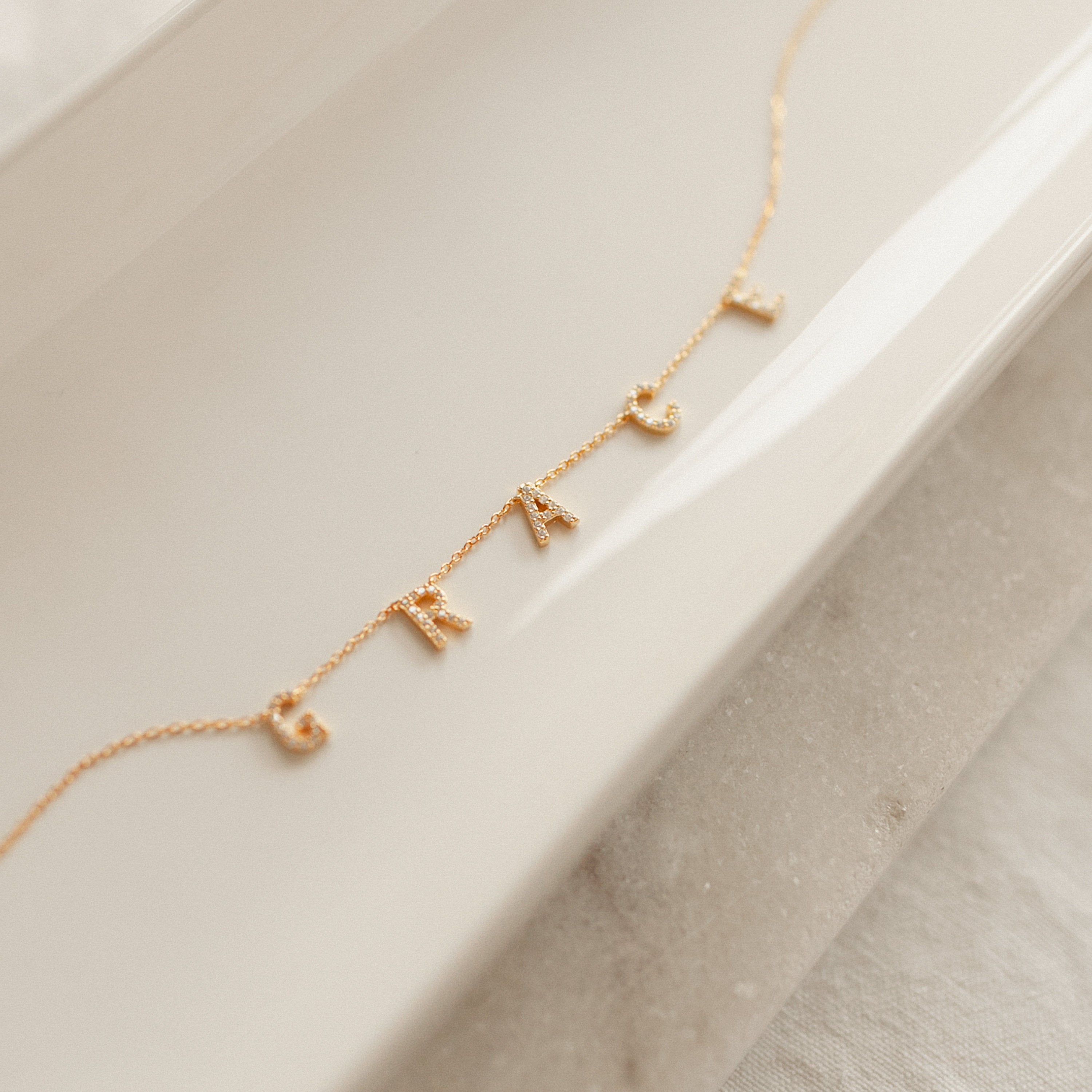 Personalized jewelry shines with this Pave Letter Necklace in Rose Gold, featuring letter charms spelling GRACE on a white ceramic dish.