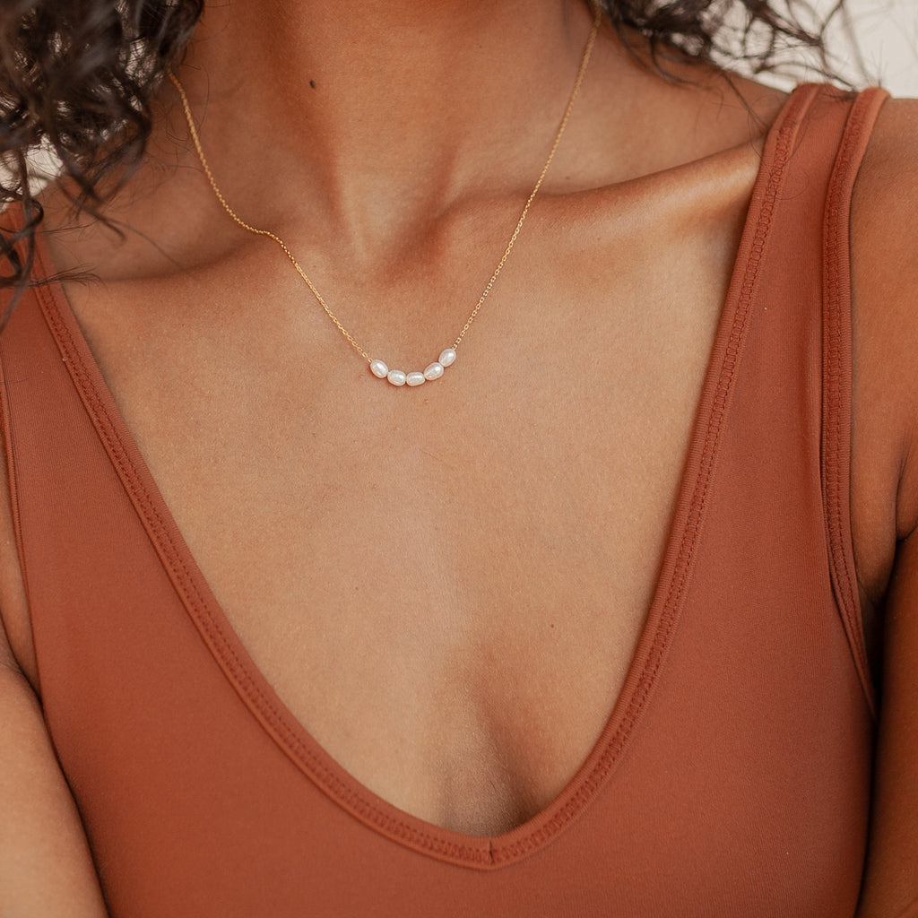 Close-up of a woman wearing a delicate gold Pearl Bead Necklace with five small white pearls arranged in a gentle curve, paired with a rust-colored V-neck top.