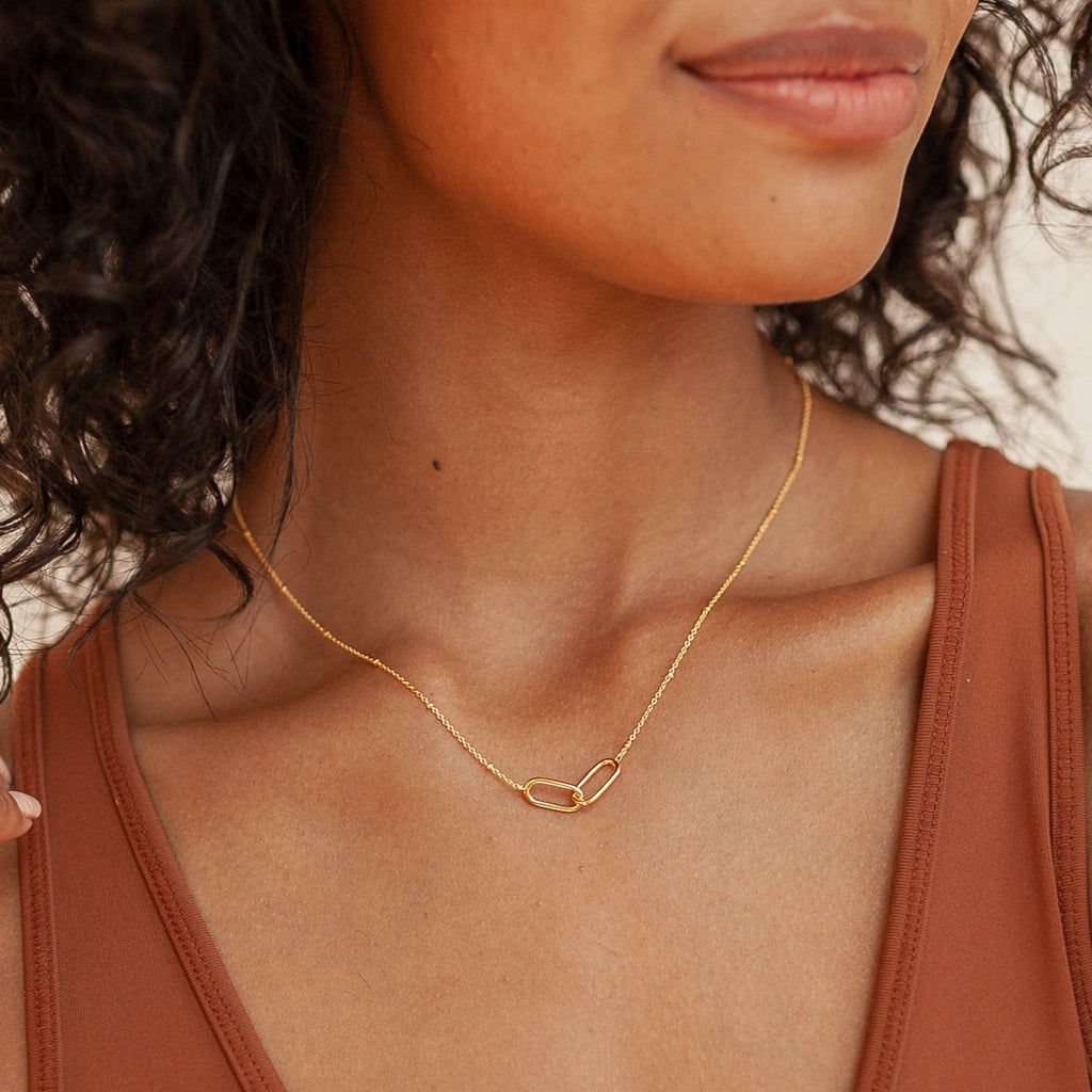 Close up of a woman wearing a delicate gold necklace featuring two interlocking oval links, styled simply against a rust colored tank top.