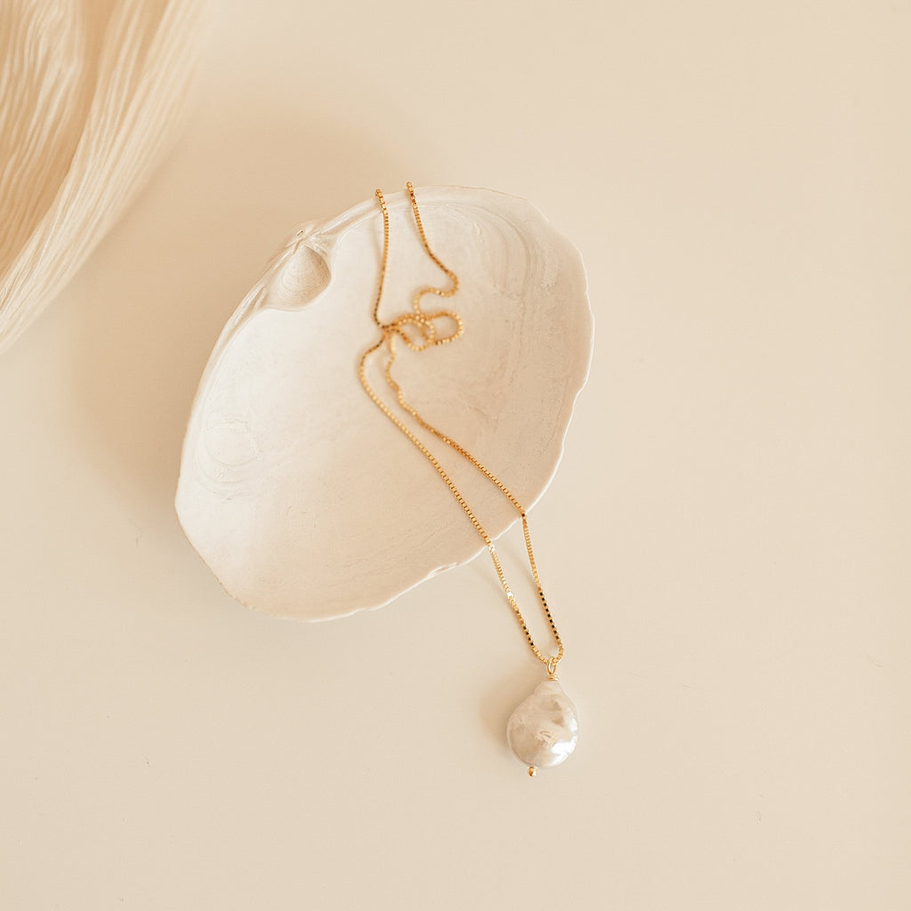 Gold chain necklace with a large, teardrop shaped baroque pearl pendant, displayed inside a textured white ceramic dish on a neutral background.