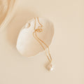 Gold chain necklace with a large, teardrop shaped baroque pearl pendant, displayed inside a textured white ceramic dish on a neutral background.