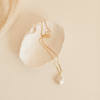 Gold chain necklace with a large, teardrop shaped baroque pearl pendant, displayed inside a textured white ceramic dish on a neutral background.