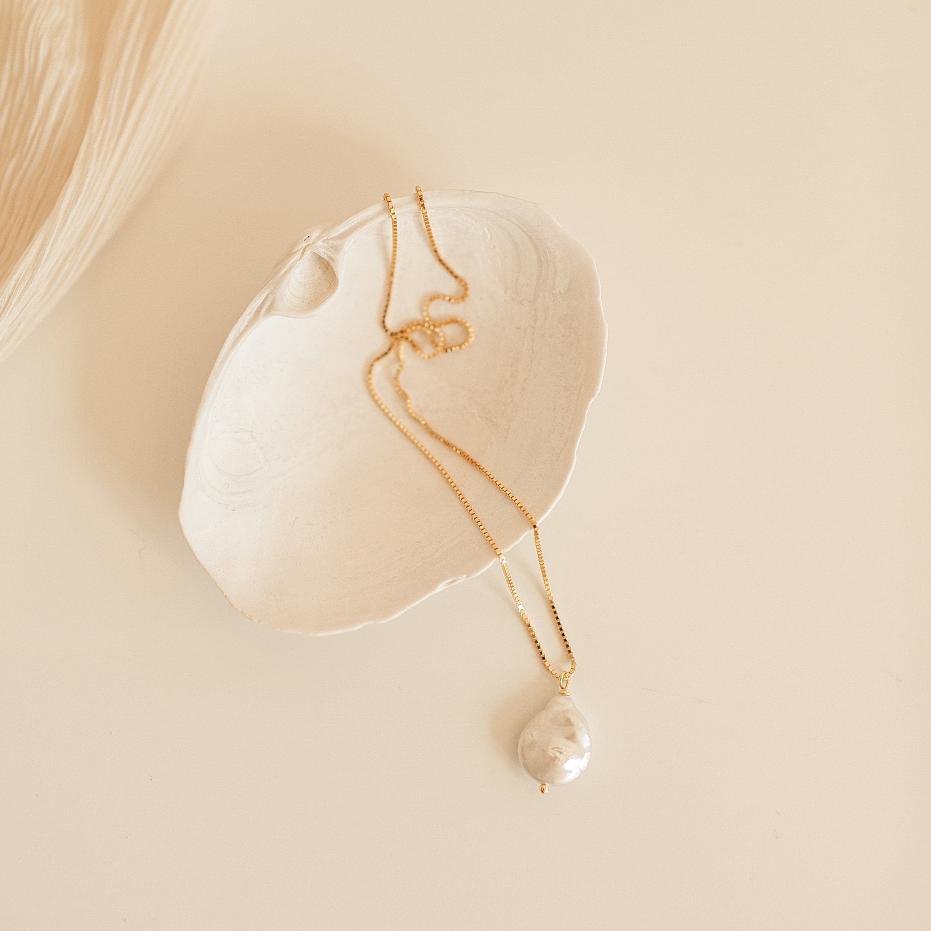 Gold chain necklace with a large, teardrop shaped baroque pearl pendant, displayed inside a textured white ceramic dish on a neutral background.