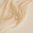 The Sadie Pearl Necklace features a round freshwater pearl pendant on a gold chain, elegantly displayed on beige fabric—an enduring choice for bridesmaids.