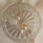 Two pairs of Baroque Pearl Earrings—one classic and one teardrop style, featuring silver and gold posts—are elegantly displayed on a clear glass dish.