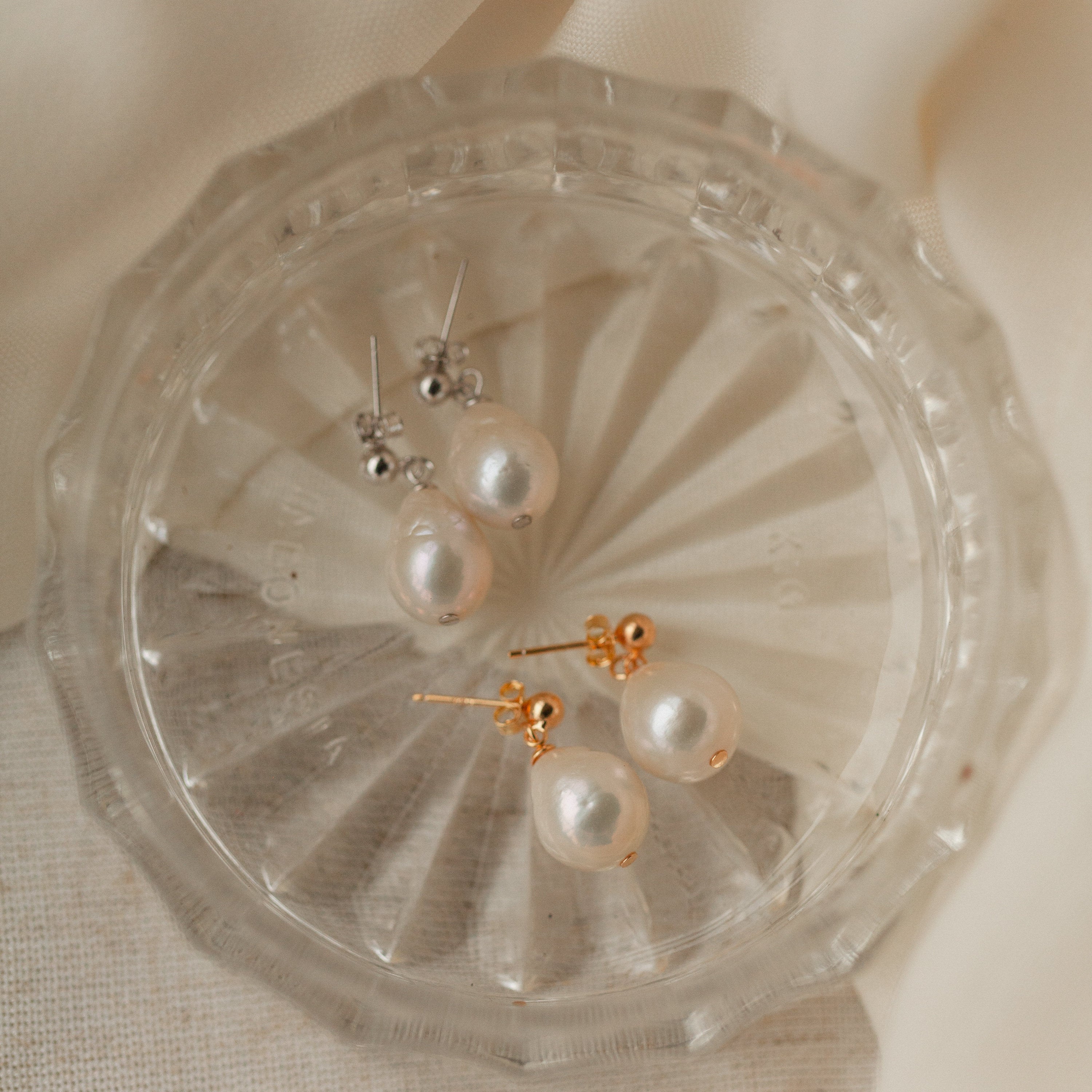 Two pairs of Baroque Pearl Earrings—one classic and one teardrop style, featuring silver and gold posts—are elegantly displayed on a clear glass dish.