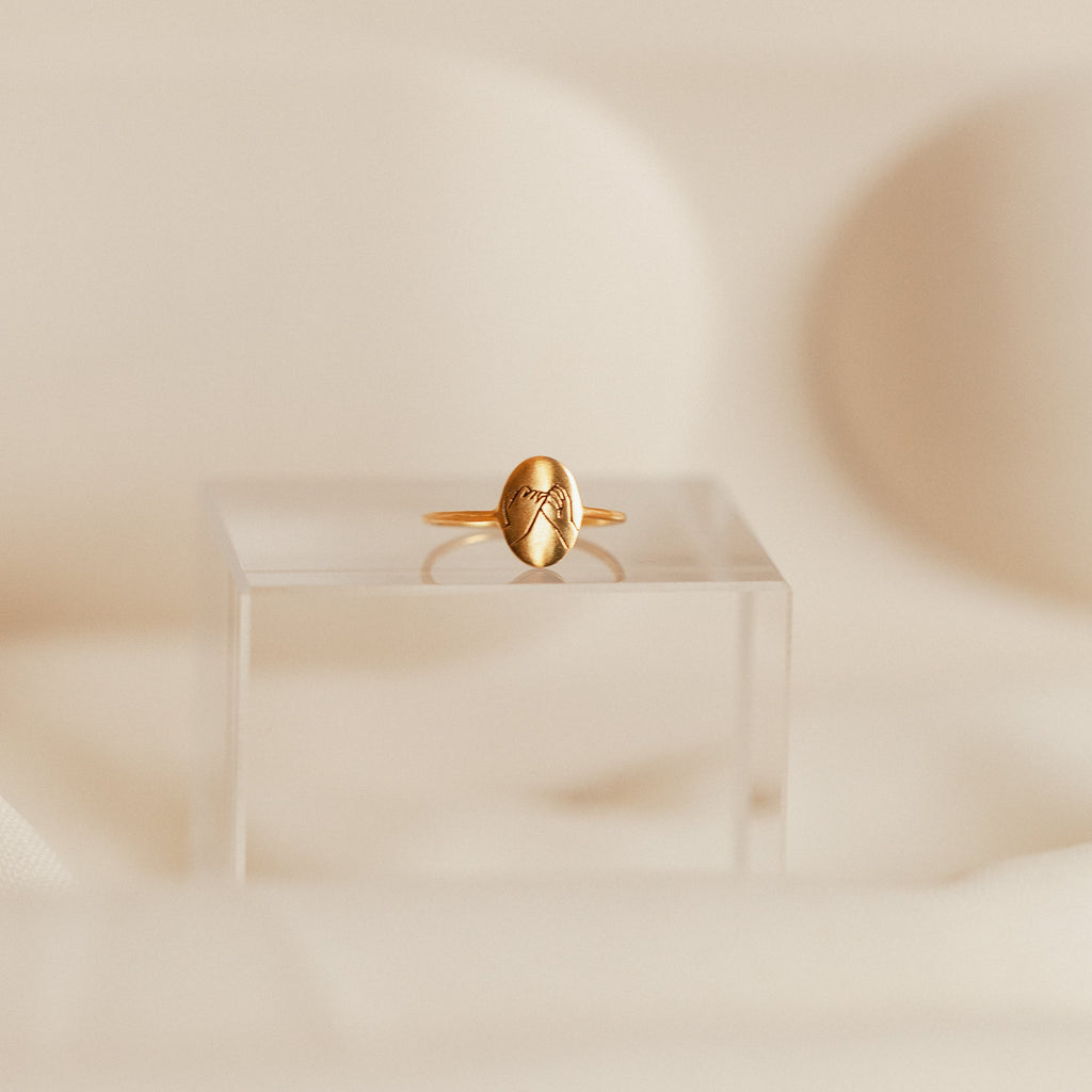 The Gestures of Love Ring features an engraved mountain design in gold, elegantly displayed on a clear acrylic stand against a soft beige background—a refined piece of figurative jewelry.