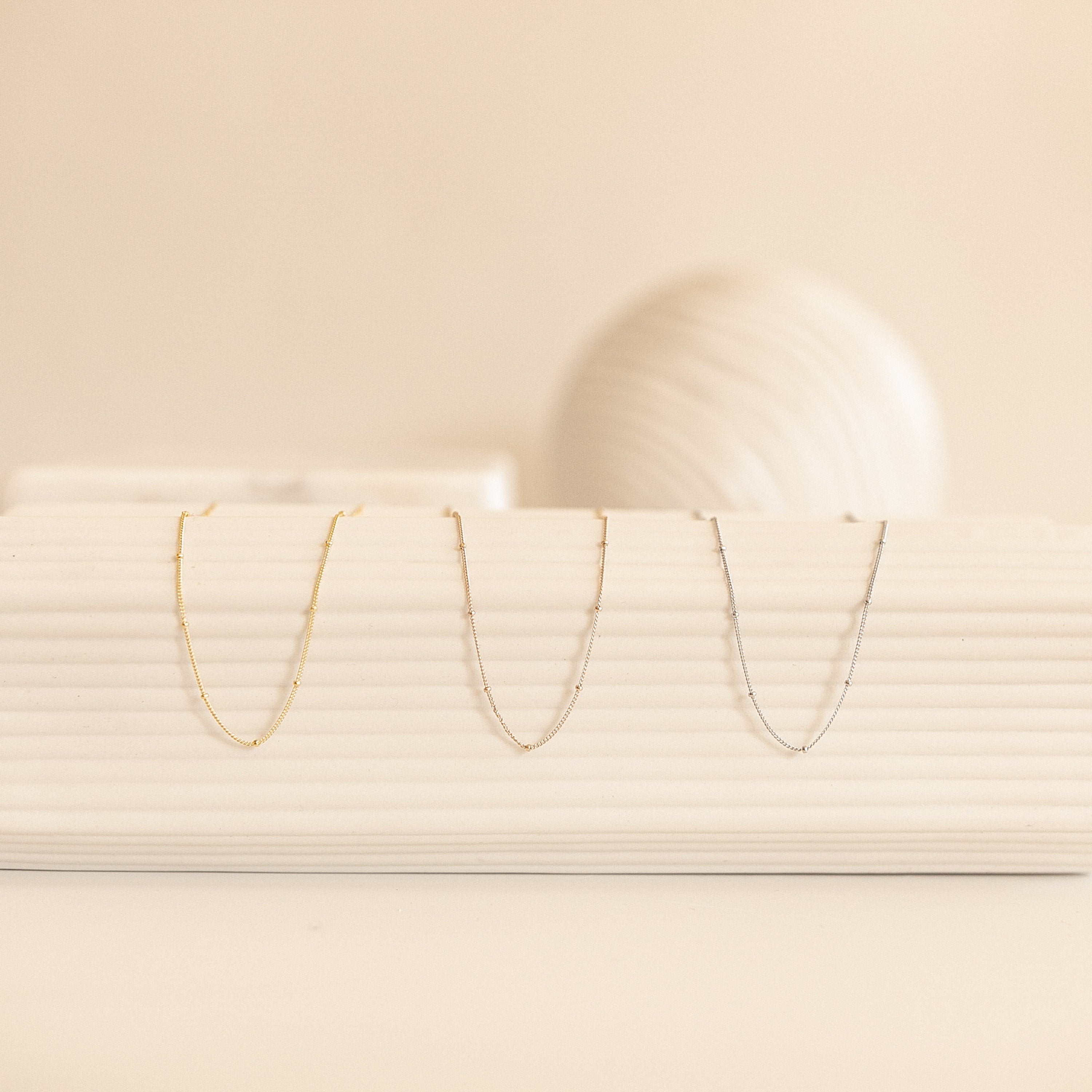 The Satellite Chain Bracelet in gold, silver, and rose gold is displayed on a ribbed white surface, pairing perfectly with delicate chain necklaces for an effortlessly elegant look.