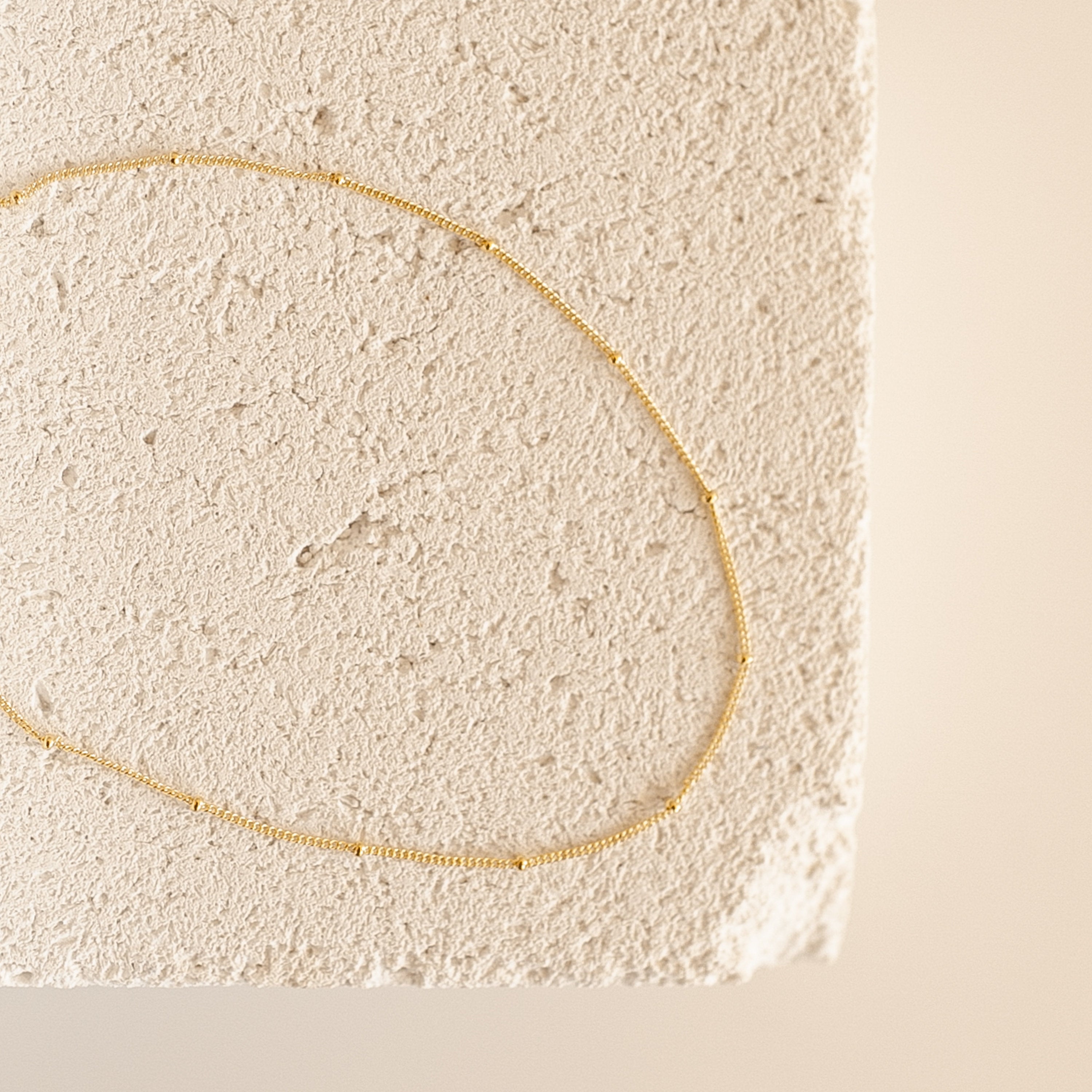 Essential Bracelet Chains with a polished gold finish are displayed on a textured, light-colored stone surface.