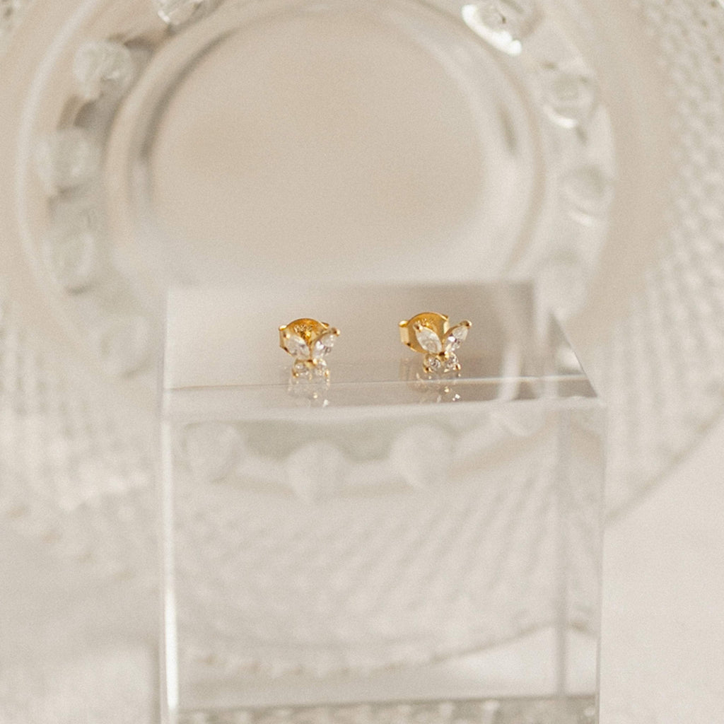 The Butterfly Diamond Studs, featuring gold and crystal with diamond accents, are displayed on a clear acrylic block against a textured background for a youthful flair.
