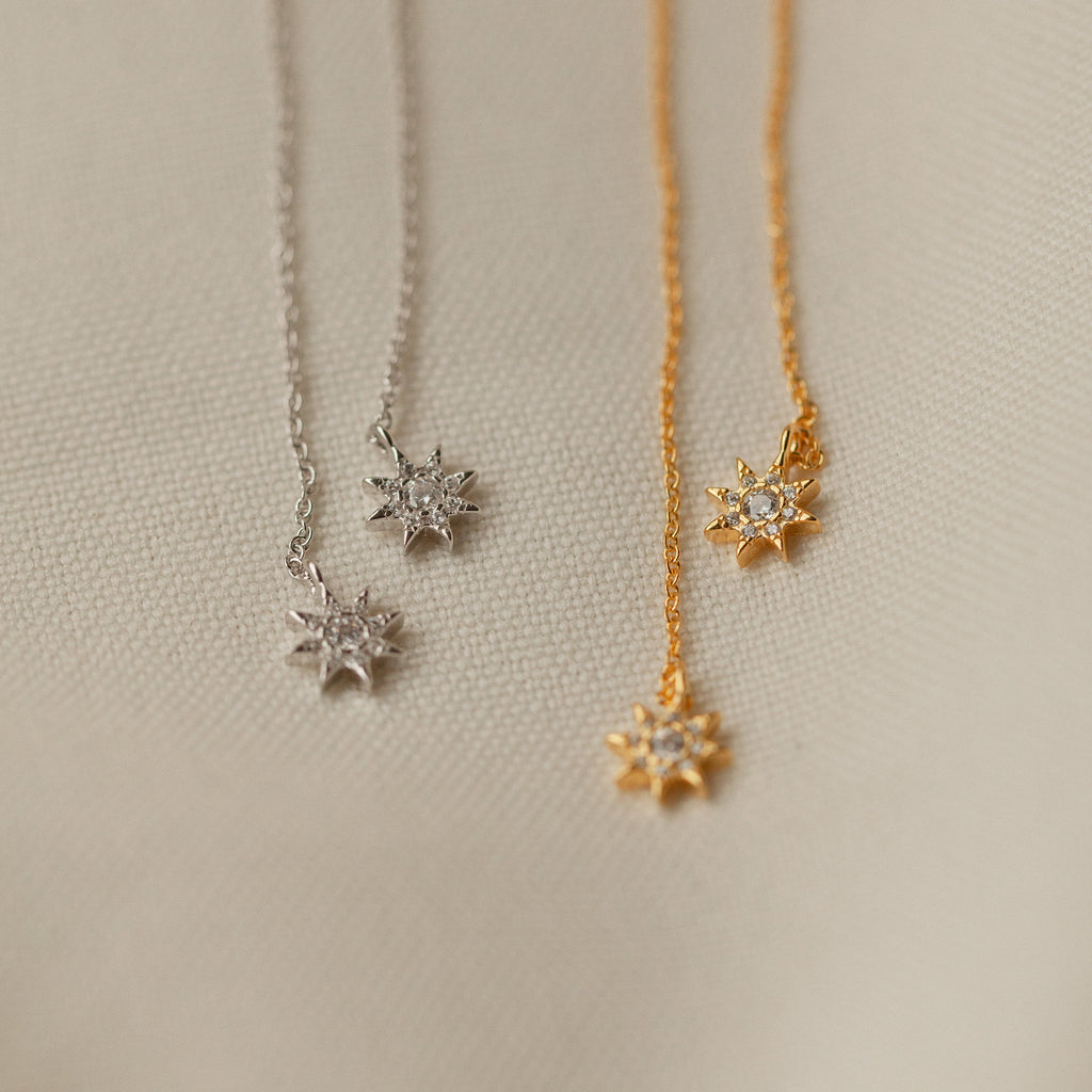 Pair these star-shaped pendant necklaces—one silver, one gold—displayed on a textured beige surface with our Lux Threader Earrings for an elegant look.