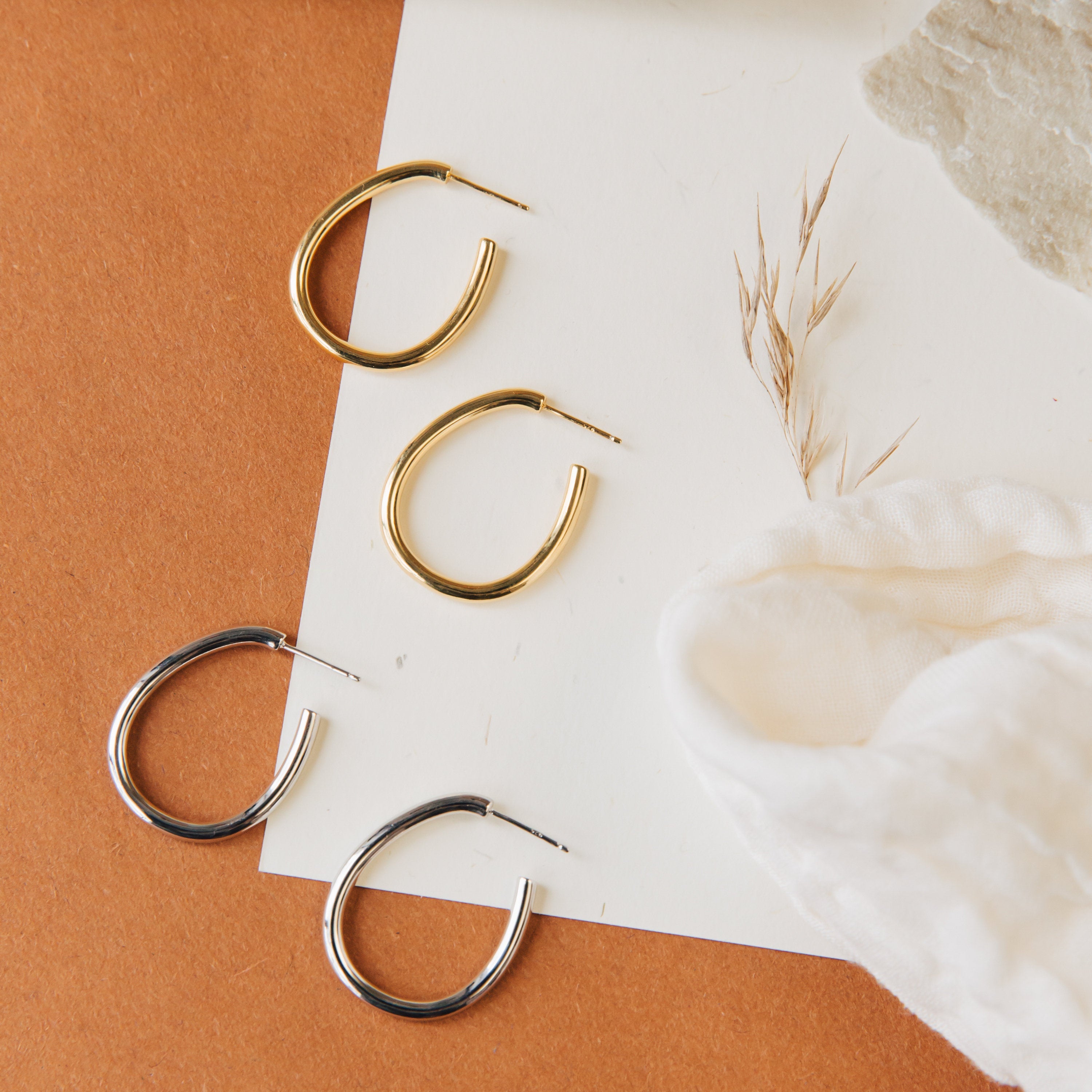 Two pairs of Anine Oval Hoops—one gold, one silver—shown on brown and white surfaces. These stylish oval hoop earrings elevate any look.