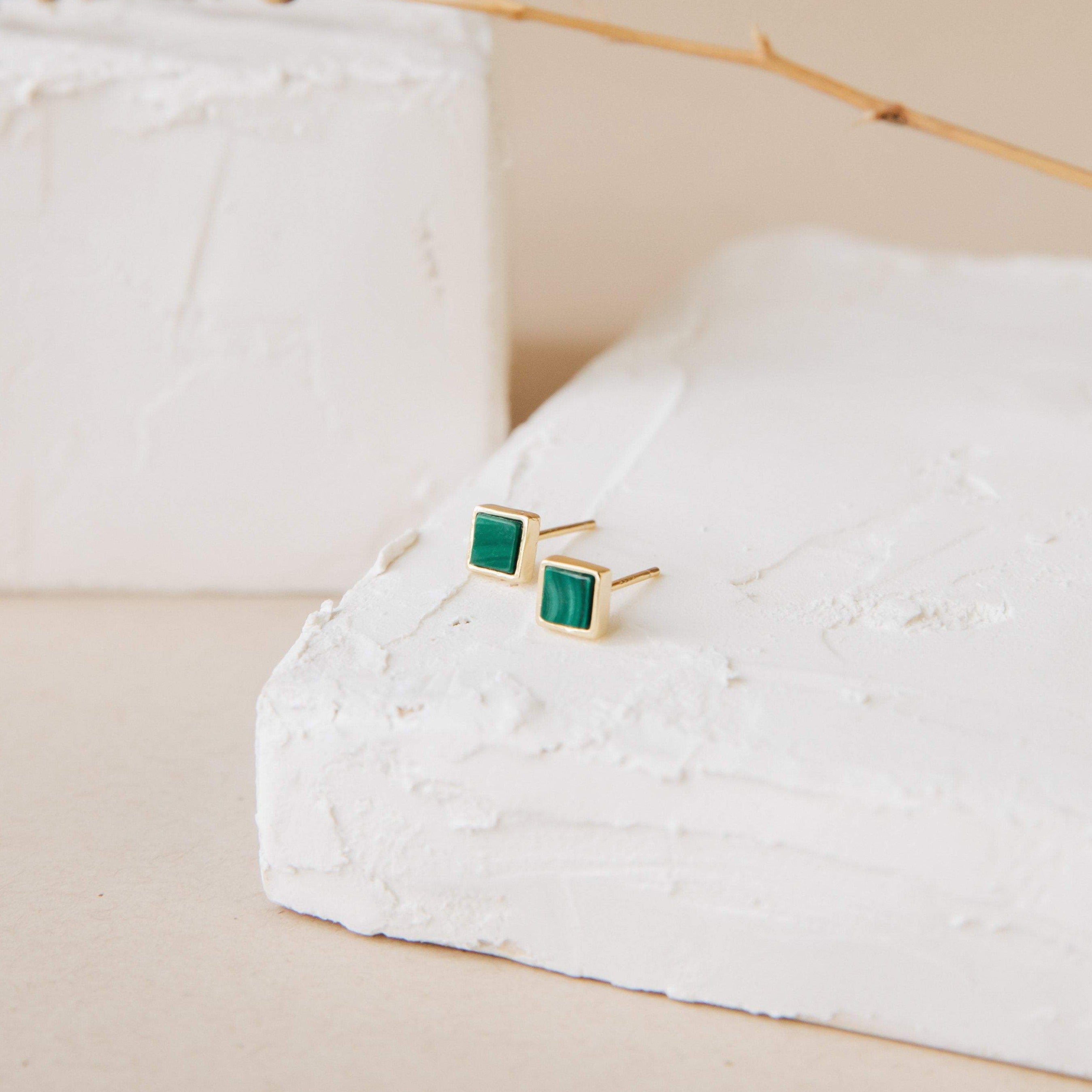Malachite Studs: Gold geometric box-shaped earrings with green malachite stones displayed on white textured blocks against a neutral backdrop.