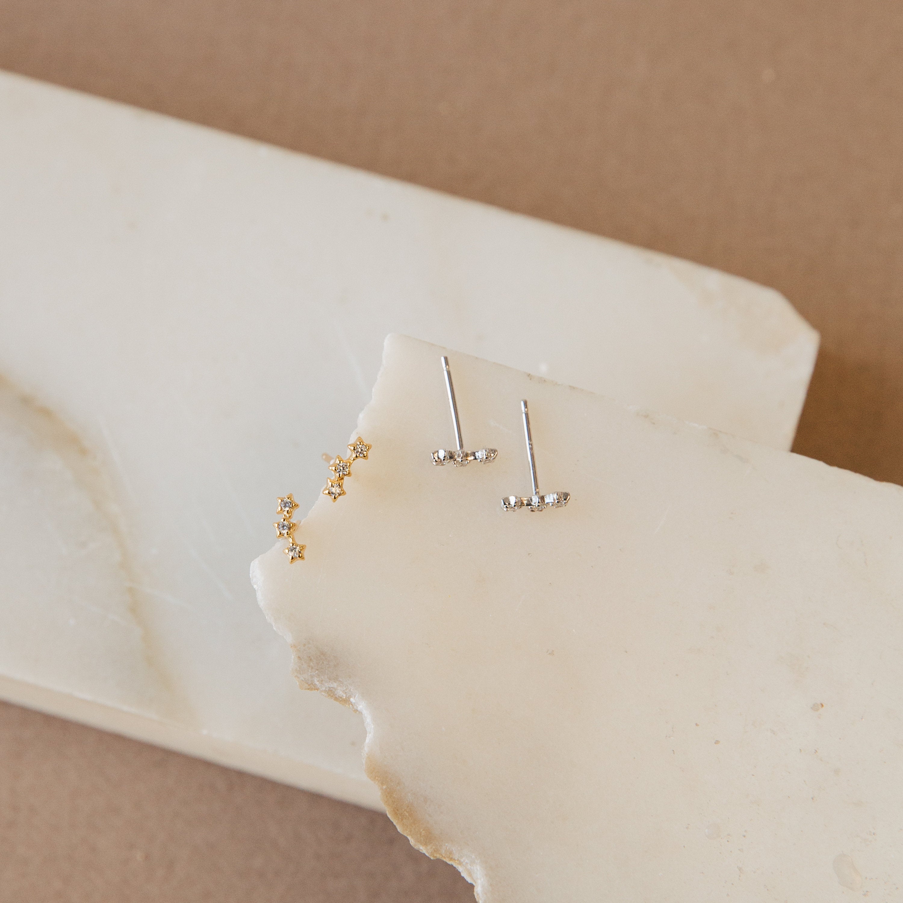 Two pairs of dainty Stella Star Studs, in gold and silver, are elegantly displayed on stacked white marble slabs.