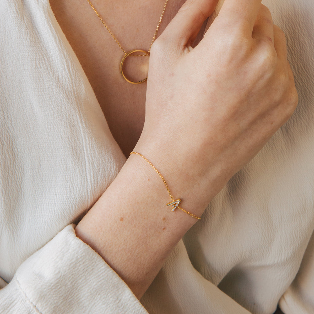 A person wears a gold necklace with a circle pendant and a Pave Initial Bracelet featuring an A charm.