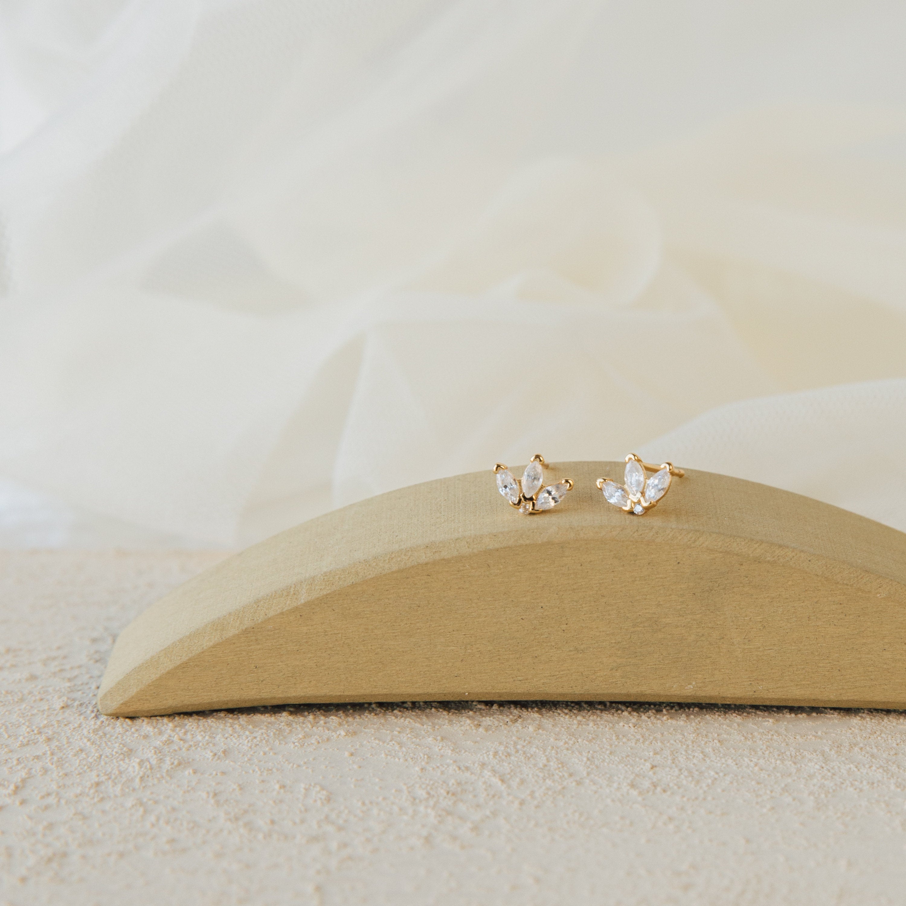 Arya Petal Studs—gold and diamond butterfly-shaped earrings—are elegantly displayed on a curved wooden stand with a soft fabric background.