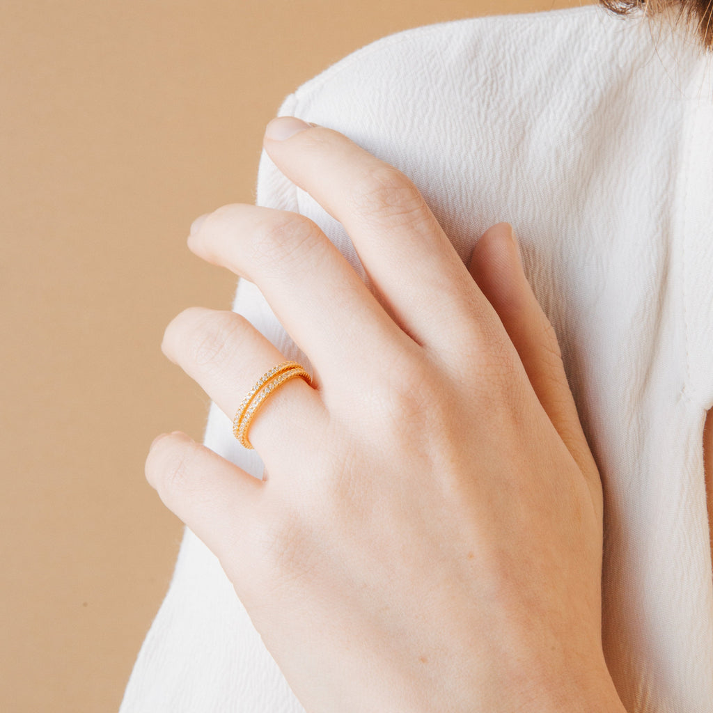 A hand wearing the Dove Eternity Ring, a petite gold band adorned with small stones, rests on a white fabric top against a beige background.