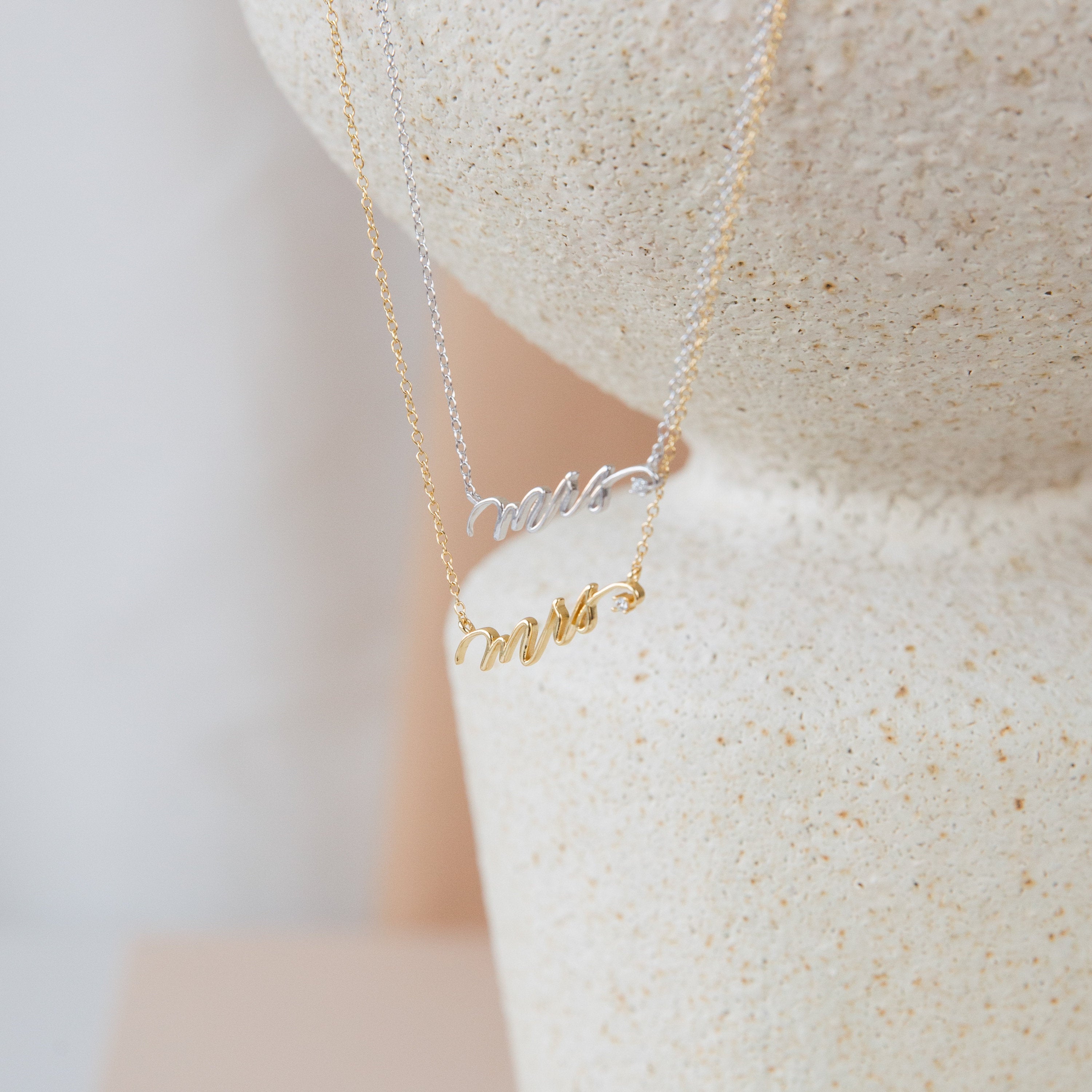The Mrs. Necklace, available in gold and 925 sterling silver, is showcased on a textured stone surface—an ideal choice for a thoughtful wedding gift.