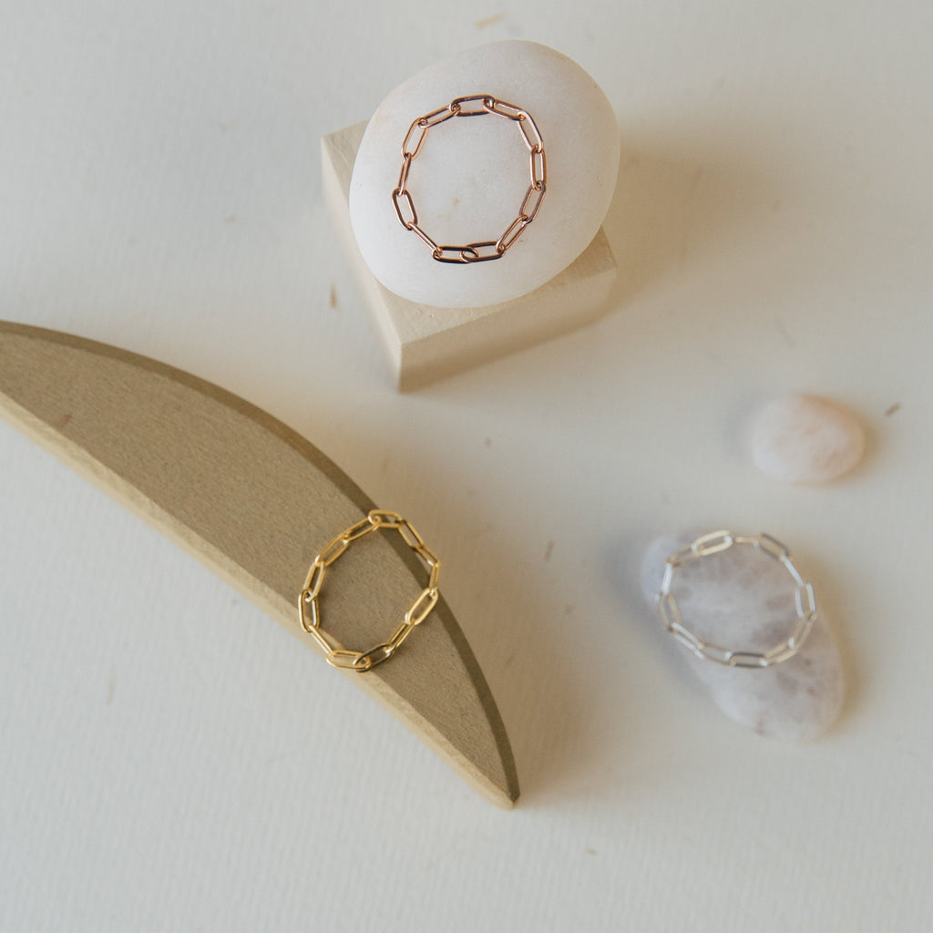 Three Paperclip Chain Rings in gold, rose gold, and silver—ideal for stacking—are displayed on stones and wooden shapes. Product: Paperclip Chain Ring.