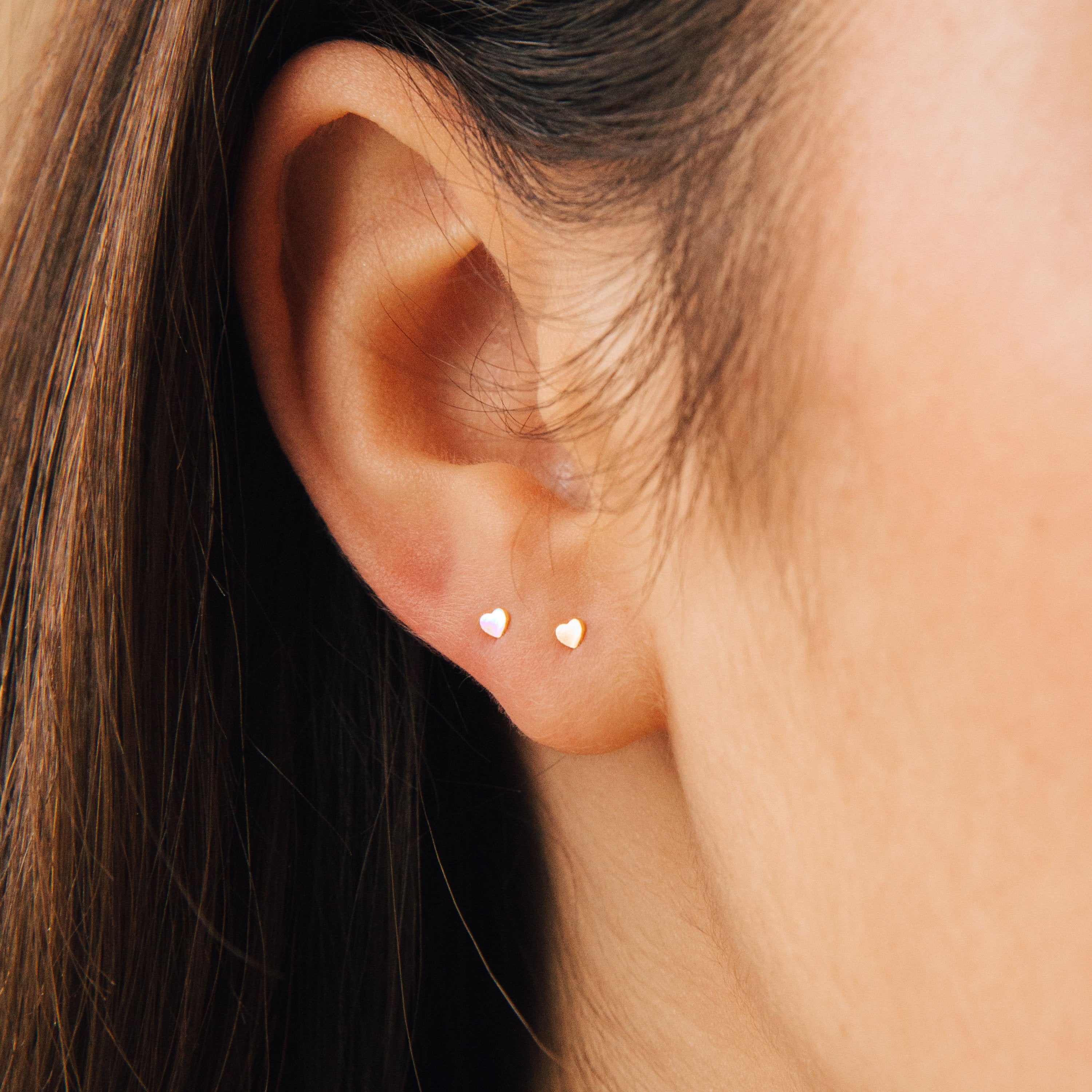 Close-up of a persons ear wearing Tiny Heart Studs in 18K Gold—ideal for daily wear or as a thoughtful gift—with brown hair softly framing the background.