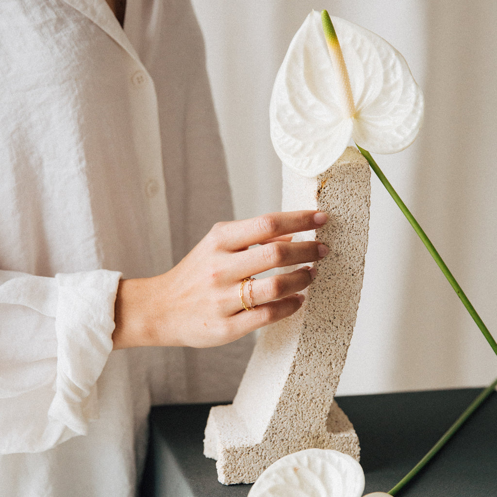 A hand in a white sleeve, wearing the Paperclip Chain Ring among stacked rings, touches a curved stone sculpture holding a white anthurium flower.