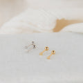 The Tiny Heart Studs in 18K Gold are displayed on a light textured surface with soft cream fabric in the background—making them a chic, minimalist gift choice.